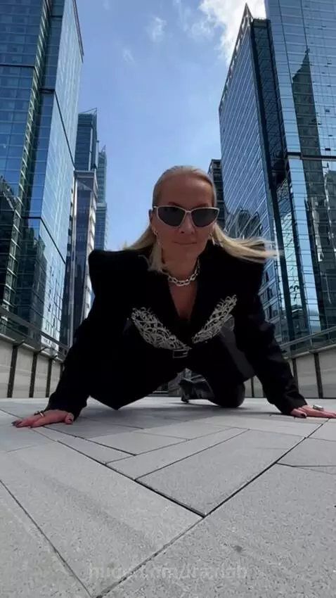Stylish woman with blonde hair in sunglasses and a black blazer with silver embroidery, posing on a city bridge.