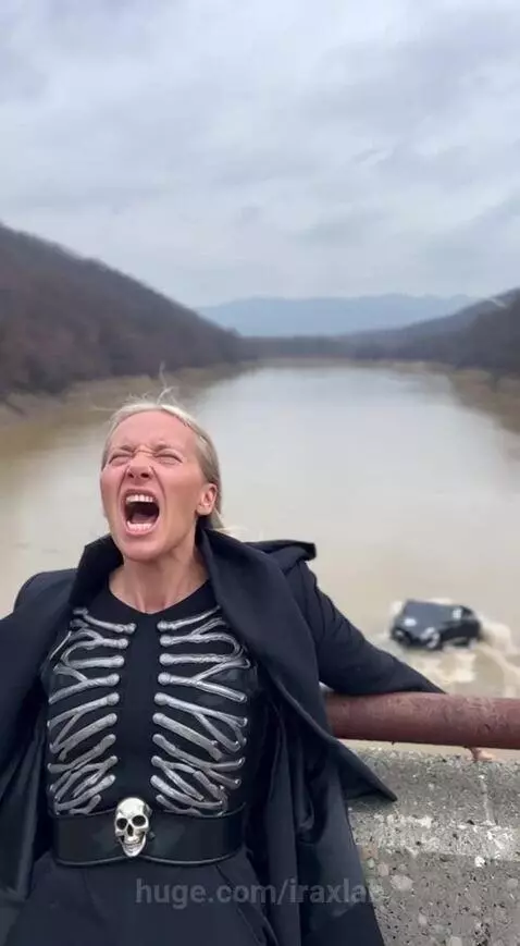 Woman in black coat and skeleton top on bridge, holding a car above her head over a river.