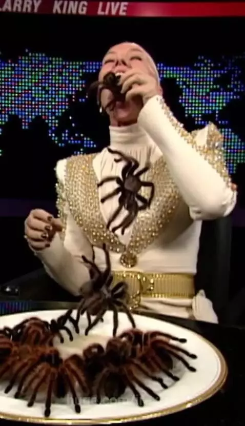 Woman in a white and gold jacket eating tarantulas at a table, with 'LARRY KING LIVE' and a world map in the background.