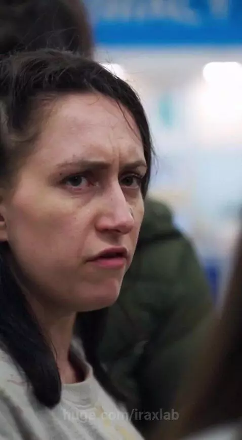 Woman in an elaborate black coat with silver studs stands in a pharmacy line, facing judgmental onlookers.