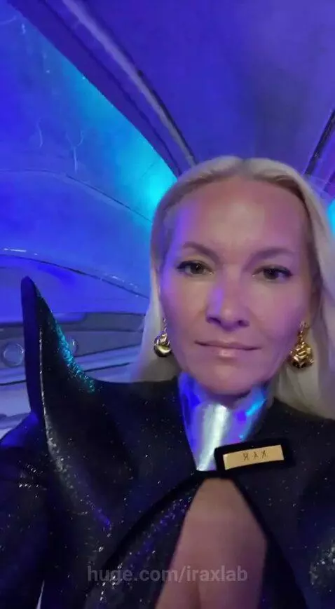 Woman in futuristic outfit inside a vehicle with blue and purple lighting, looking out at UFOs in a cloudy sky.