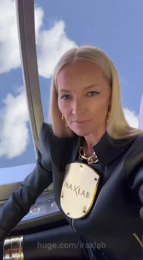 Woman in leather jacket and gold chain, seated in a futuristic vehicle, looking at camera, clouds visible through window.