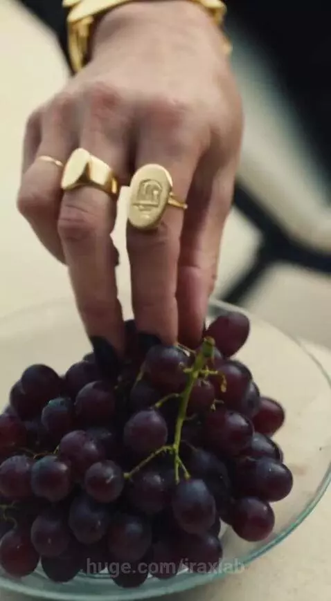 Stylish woman with blonde hair in black outfit eating red grapes from a glass bowl, gold jewelry, man in background.