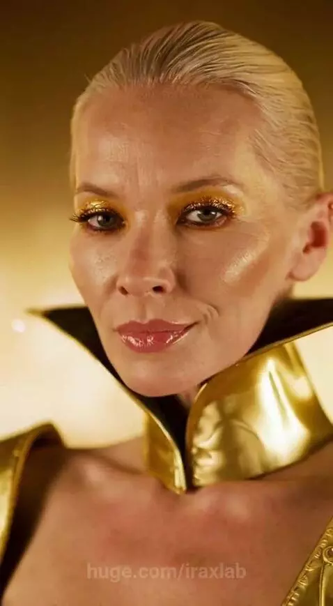 Woman with blonde hair and gold glitter eyeshadow in a form-fitting golden dress with a cape, standing against a shimmering gold wall.
