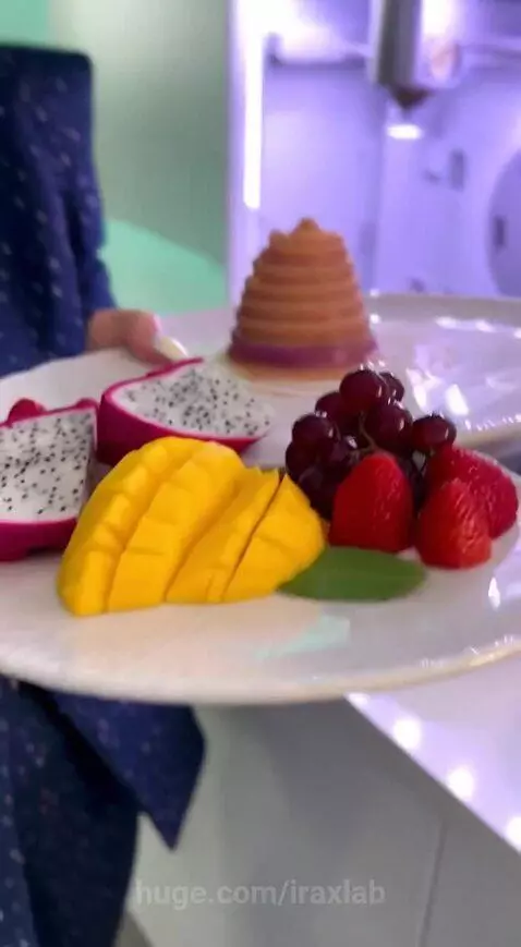 Woman in blue suit observing a 3D food printer creating a layered dessert, later tasting it.