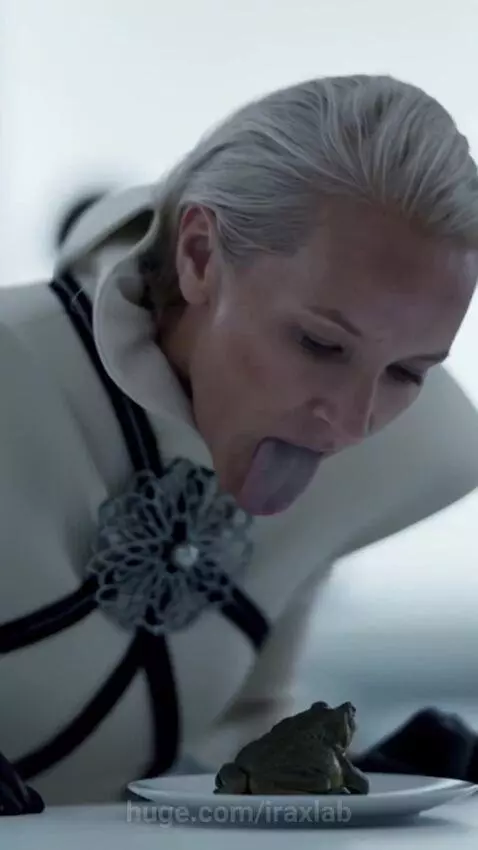 Woman in futuristic attire licking a toad on a plate in a brightly lit room with other people observing.