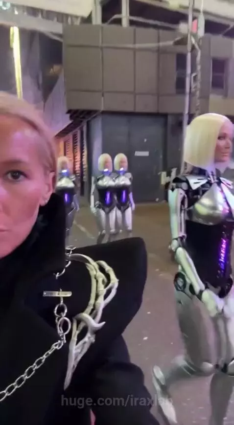 Woman filming uncontrolled humanoid robots with glowing purple lights on a city street at night.