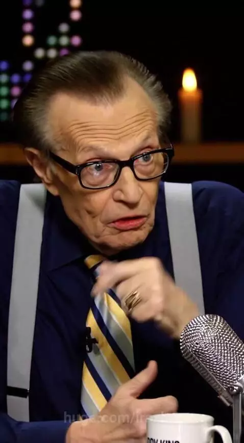 Woman dressed as a vampire with pale skin, fangs, and yellow eyes, wearing a black and gold outfit, sips red liquid.