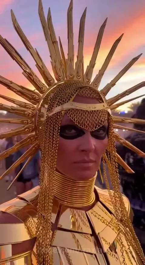 Woman in elaborate golden costume with spiked headdress and armor at Coachella festival at sunset.