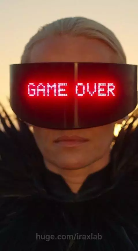 Futuristic person in a black and silver outfit with a feathered collar and 'GAME OVER' visor walks across desert dunes at sunset.