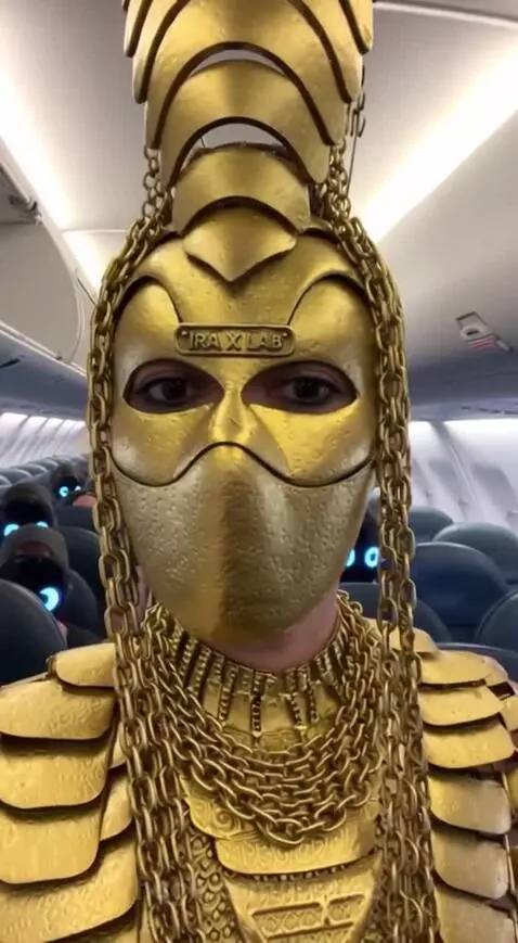 Person in elaborate gold costume in an airplane aisle with identical passengers wearing masks with glowing blue eyes.