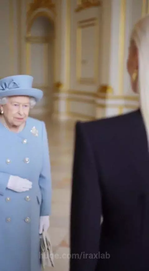 Queen Elizabeth II in a blue coat and hat, signing a card with a gold pen, held by a woman in a black blazer.