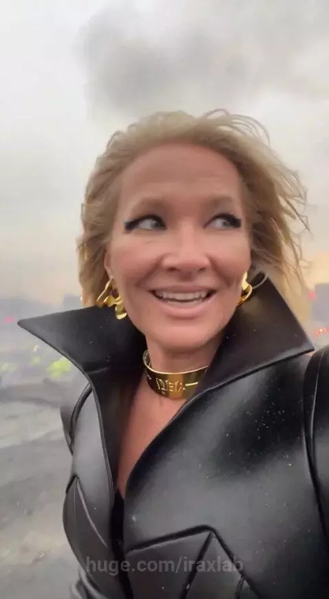 Blonde woman in black coat with gold accessories, recording herself with fires and smoke in the background at sunset.