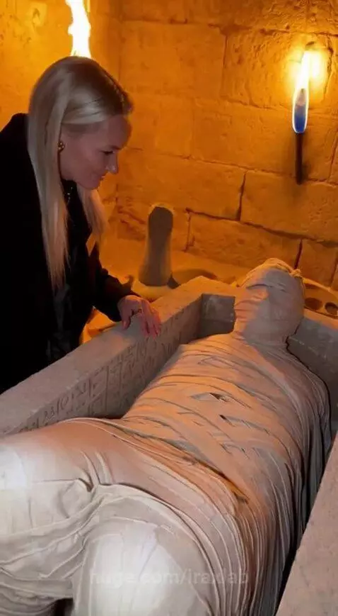 Woman in black outfit startled by a mummy's moving leg inside a sarcophagus in an ancient tomb.