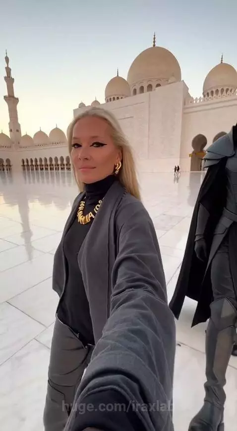 Woman in gold necklace and black turtleneck walks in a white marble courtyard with a mosque in the background, beside a fantasy-armored figure.