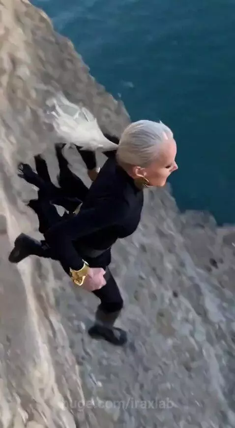 Woman with ponytail and gold accessories jumps off a cliff towards an ocean view, landing on a large inflatable pad below.