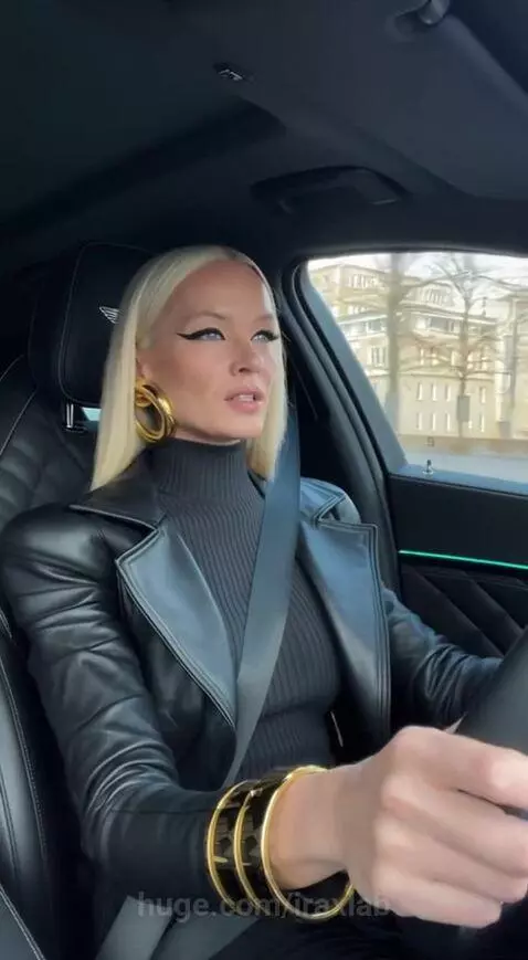 Blonde woman with winged eyeliner and gold jewelry driving a luxury car with quilted interior.