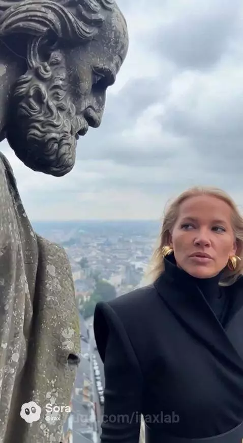 Woman in black coat with gold belt on building edge talks to stone statue, city below.