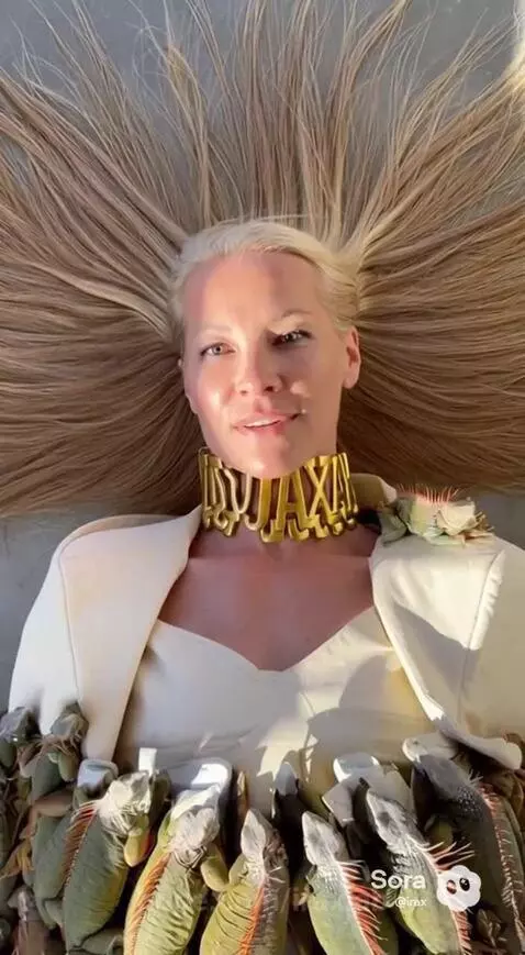 Woman lying down covered in many live green iguanas from the waist down, smiling.