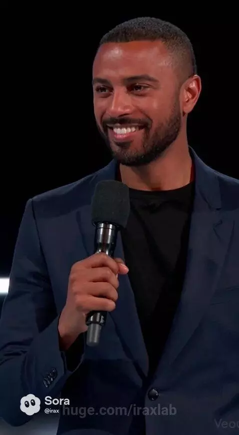 Man interviews a humanoid robot with a blonde wig on a stage, audience applauds.