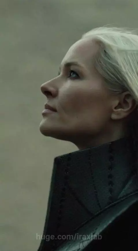 Blonde woman in futuristic black coat with gold accents stands in a sci-fi landscape with a giant robot.