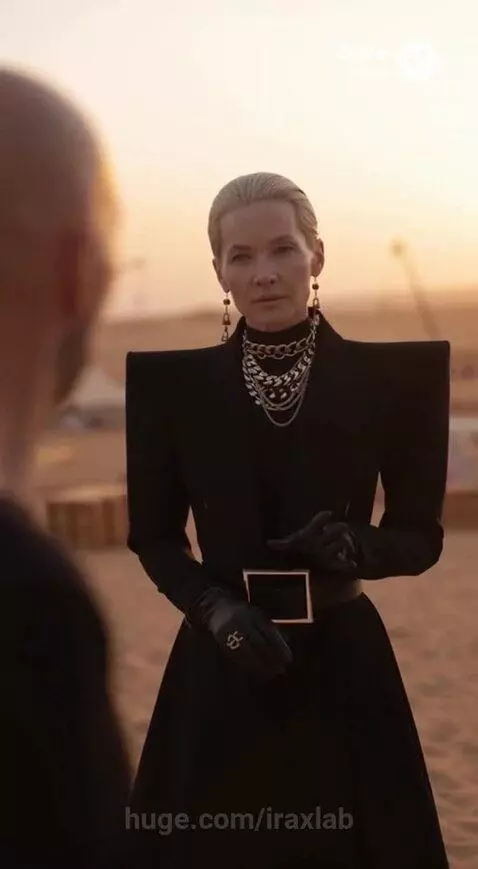 Woman in black outfit with shoulder pads and chains in desert at sunset, disappearing in light.