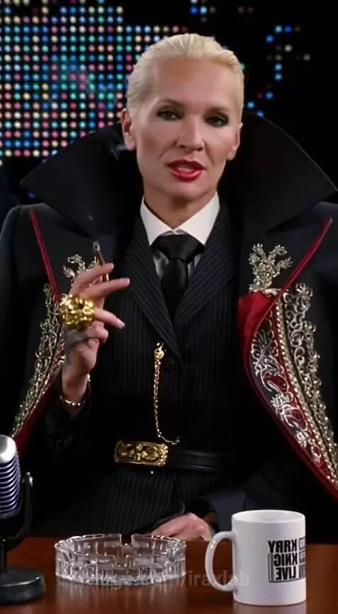 Woman in a pinstripe suit and elaborate coat holding a cigar, sitting at a desk with a microphone.