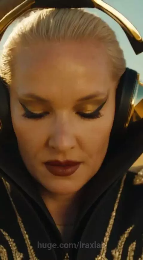 Woman with blonde hair and gold headphones DJing in the desert, with large metallic snake sculptures in the background.