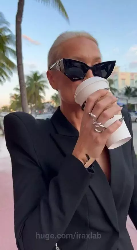 Woman in black blazer and patterned skirt sips coffee on Ocean Drive with palm trees and Art Deco buildings.