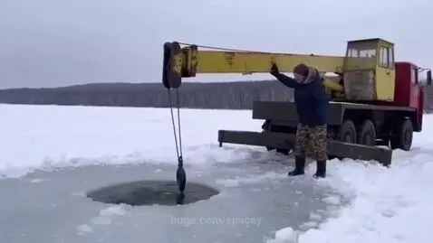 Man in winter clothes on a frozen lake next to a crane pulling a large, dark, whale-shaped object from an ice hole.