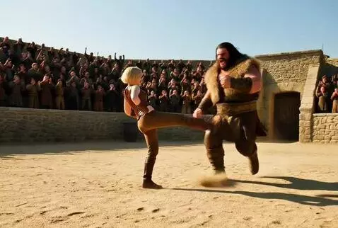 Blonde woman in a brown outfit stands triumphantly over a fallen barbarian in a sandy arena, smiling at the crowd.