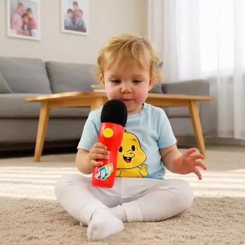 A young child with blonde hair sits cross-legged, holding a red toy microphone and making expressive faces while singing.