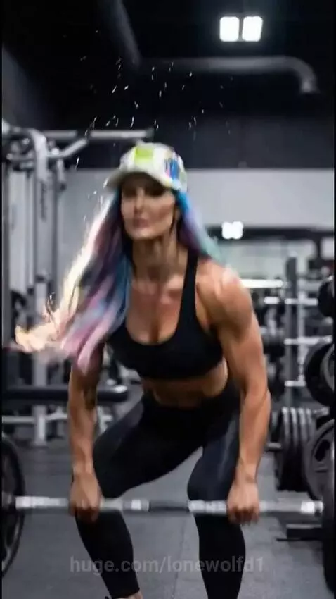 Muscular woman with multi-colored hair in a baseball cap performing a deadlift in a gym.