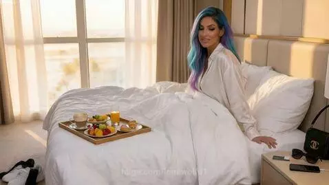 Woman with blue and purple hair in a white satin shirt, smiling with a breakfast tray on the bed in a hotel room.