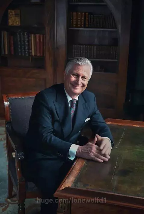 A distinguished man with grey hair and a suit smiles warmly at a wooden desk, preparing a royal march.