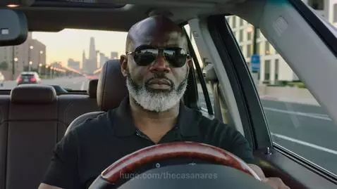 Bald Black man in his 60s with salt and pepper beard and sunglasses driving a luxury SUV at dawn in the city.
