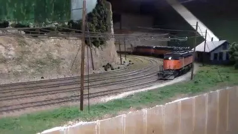 HO scale model train layout with Milwaukee Road E77 electric locomotive pulling freight cars past a miniature house and hillside.