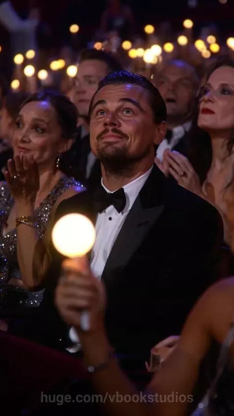 Leonardo DiCaprio in a tuxedo smiling and clapping at a formal event with glowing lights.