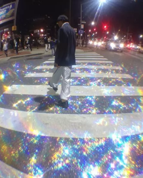 Person walking across a crosswalk at night illuminated by vibrant, sparkling rainbow lights.