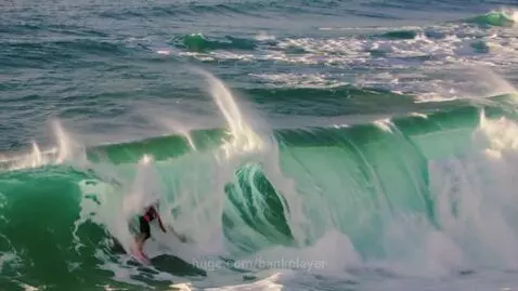 Surfer in dark attire on a pink surfboard riding inside a massive, breaking turquoise ocean wave barrel.