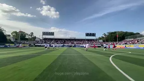 Wide-angle view of a soccer match on a sunny day in a stadium with players on the field and spectators in the stands.