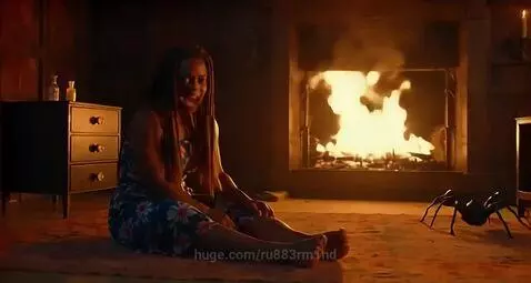 Woman with braids sitting by a fireplace, facing a spider-like object, looking determined.