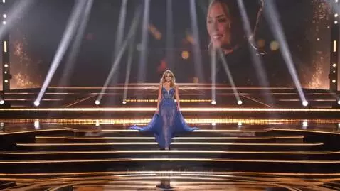 Woman in a sparkling royal blue evening gown with a flowing train, posing on an illuminated stage during a beauty pageant.