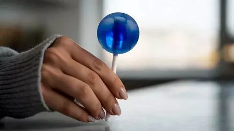 Woman with black hair and red streak in a gray cardigan and denim shorts, barefoot, holding a blue lollipop in a loft apartment.