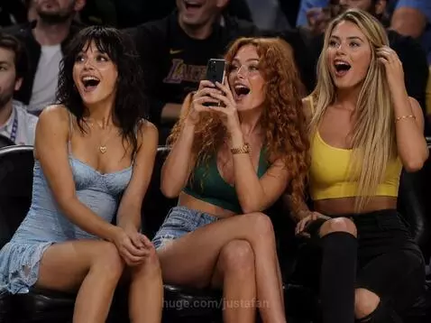 Three young women in stylish outfits react with excitement and smiles while sitting courtside at a basketball game.