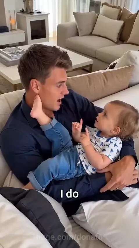 A baby sits on a man's lap on a couch, looking up at him as they talk.