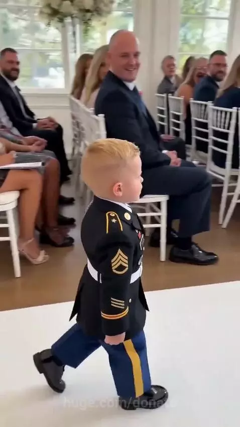 A young boy in a military uniform saluting as he walks down a wedding aisle with a ring pillow.