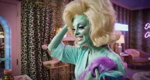 Humanoid aliens with green skin and blonde wigs, and aliens in metallic suits by a pool in a desert landscape.