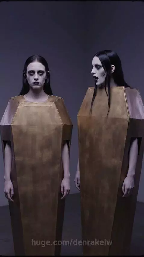 Two women in gothic makeup and coffin costumes, one revealing a blue latex suit after opening her coffin.