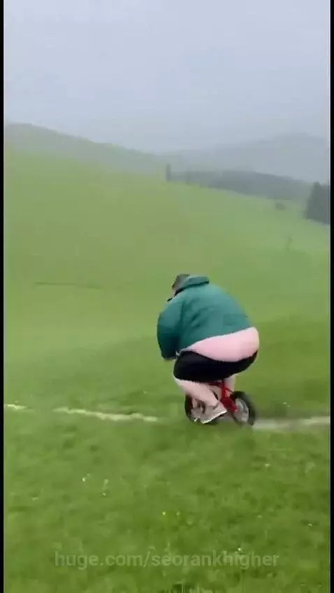 Man in green jacket and black shorts falls off a small red bicycle after attempting a dirt ramp jump.